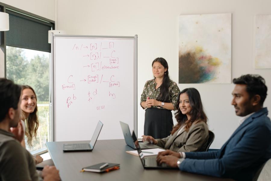 A group of students at Texas Woman's University (TWU) are in a classroom. One student stands near a whiteboard filled with mathematical notations, presenting to peers seated around a table with open laptops, attentively listening.