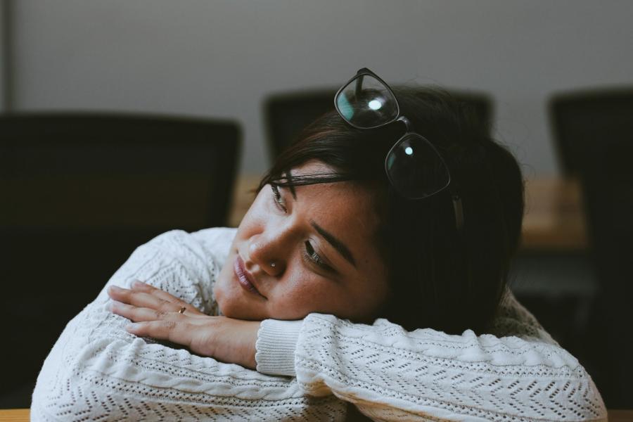 A young woman in a white knitted sweater resting their head on crossed arms at a wooden table.