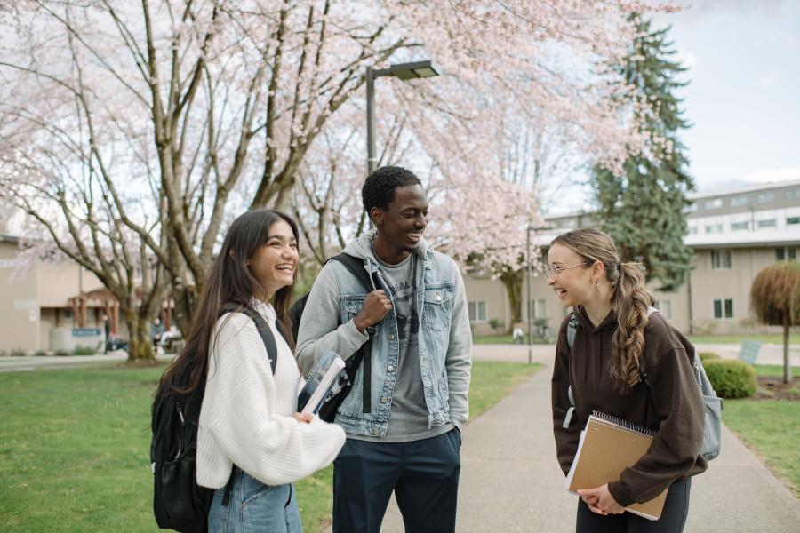 Three students standing outdoors on a university campus, holding books and backpacks, with cherry blossom trees in the background.