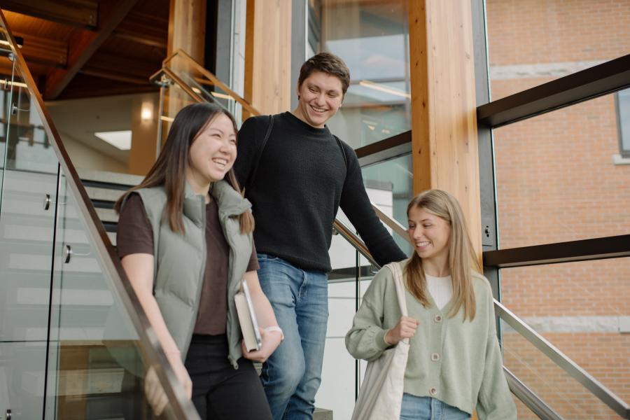 Three university students walk down a staircase inside a building with large windows and wooden beams.