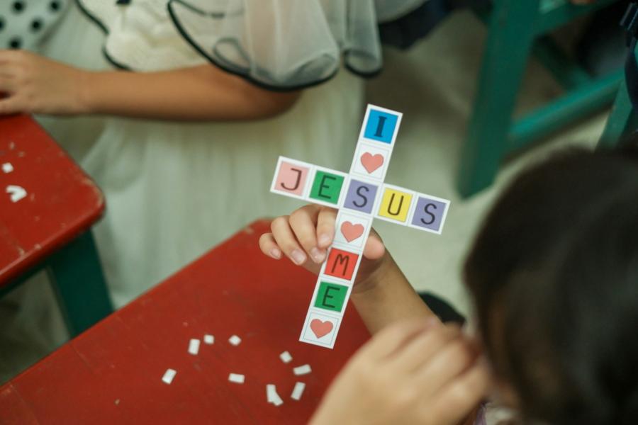Child holding a cross-shaped craft made of letter tiles spelling 'I ♥ JESUS ♥ ME' at a red table.
