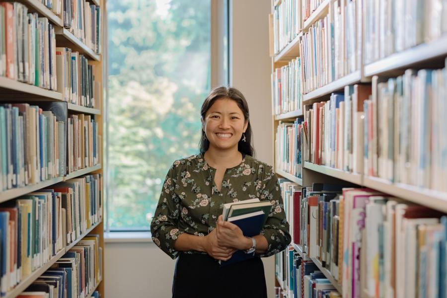 A student smiling at the camera while standing between bookshelves in a library.