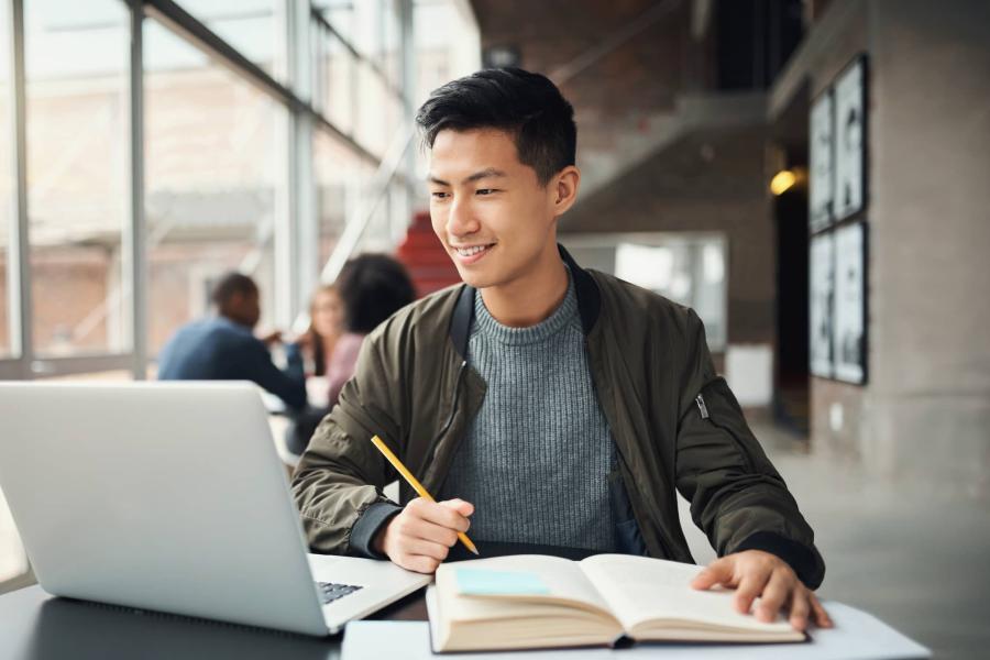 image of a person looking at the laptop, holding a pen and has a book infront of him 