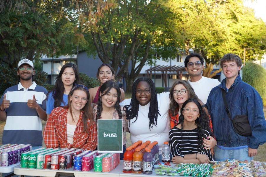 Student representatives from TWUSA pose behind a table stocked with drinks and snacks during an outdoor campus event on TWU's Langley campus, promoting student engagement and community involvement.