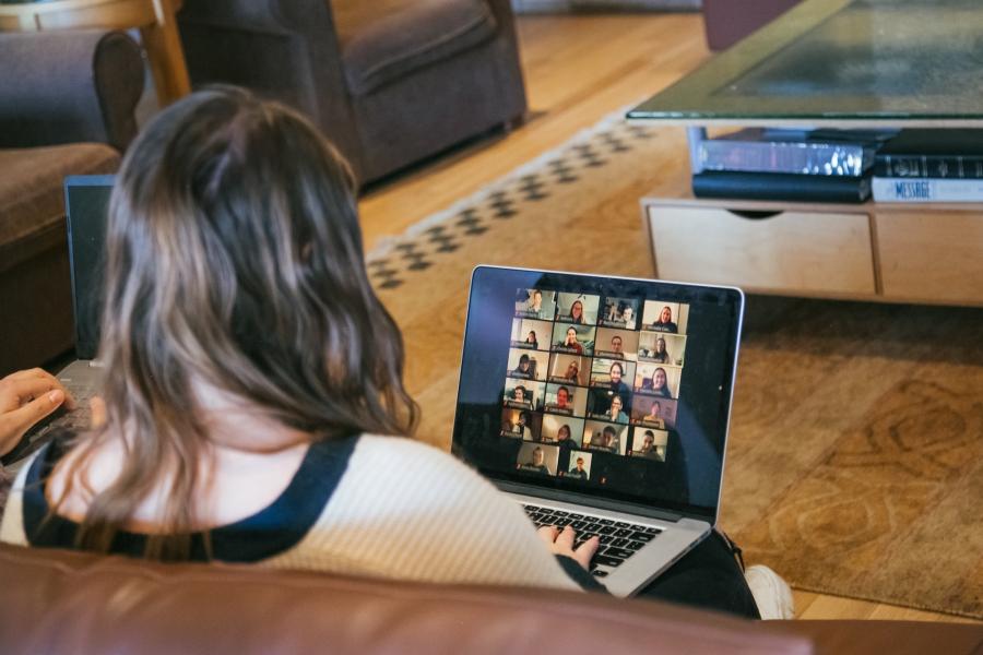 Person seated in a living room using a laptop to participate in an online video meeting, with multiple participants displayed in a grid on the screen. Another laptop is visible nearby, and the space includes comfortable seating, a coffee table, and a rug.