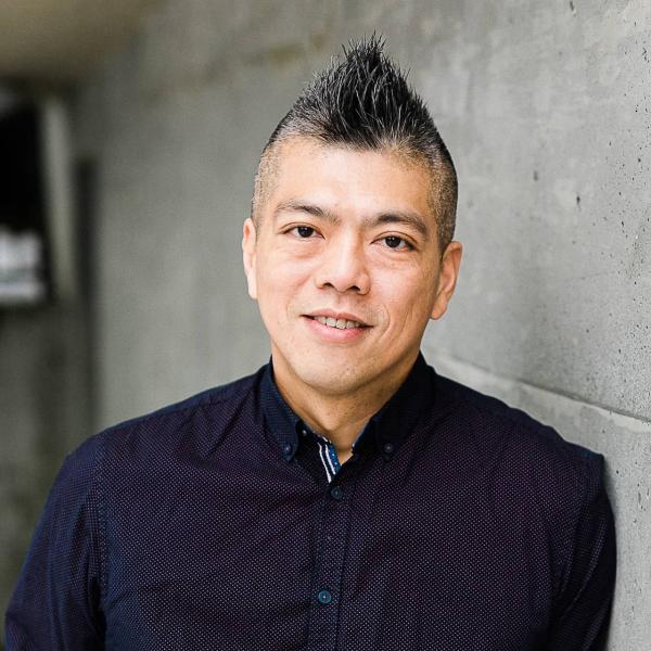 A man stands against a concrete wall, wearing a dark-colored button-up shirt with a subtle pattern.