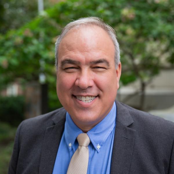 John Waind with short, light-colored hair is wearing a blue dress shirt, a beige tie, and a dark gray blazer. The background shows greenery and part of a building.