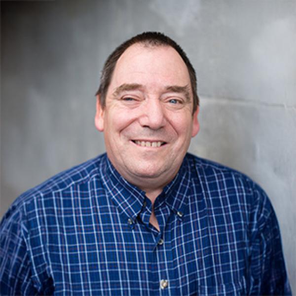 Gordon Moulden in a blue checked shirt against a gray wall.