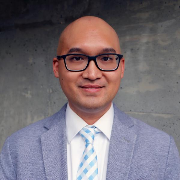 Bald man in glasses wearing a gray suit and blue tie, standing against a concrete wall.