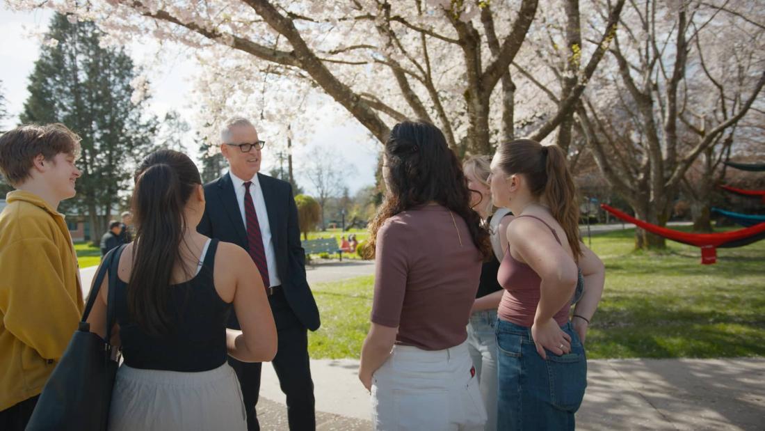 Dr. Todd F. Martin, wearing a dark suit and red tie, is talking to a group of students at Trinity Western University in Langley, BC. The students are standing in a semi-circle around Dr. Martin, engaged in conversation. The background features blooming cherry blossom trees and a sunny day with clear skies. There are colorful hammocks tied between the trees, and other people can be seen enjoying the outdoor space.