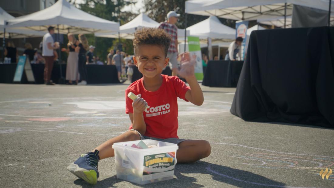 Child in a red 'Costco' shirt sitting on the ground at an outdoor event, holding chalk with a container of colourful chalks nearby and waving at the camera.