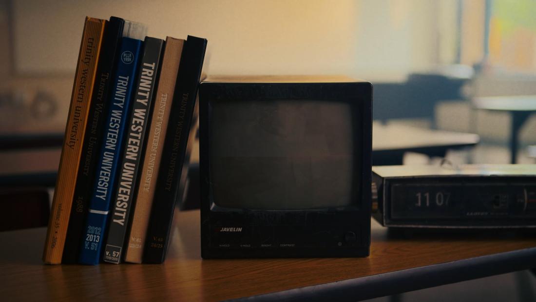 Trinity Western University yearbooks beside a vintage television and digital clock on a wooden desk.