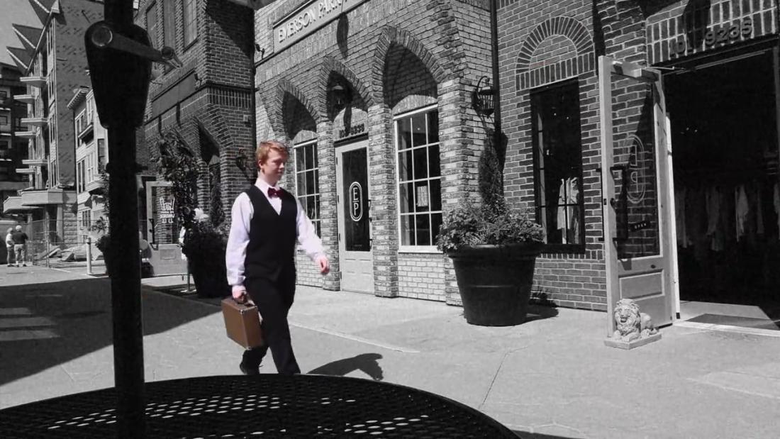 Person carrying a small briefcase walks along a sidewalk in front of brick storefronts on a sunny urban street. The image is mostly black and white, with the person shown in colour.