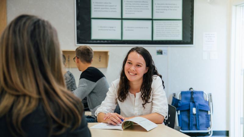 Female student at a desk, smiling at her classmate, with a booklet open.