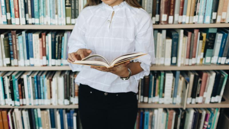 Student holding an open book in the library in front  of a shelf of books.