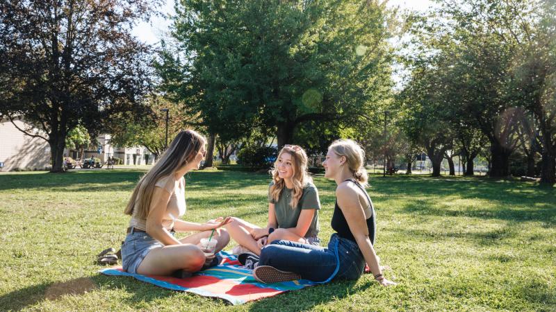Three female students sitting on a blanket in the grass on campus talking.