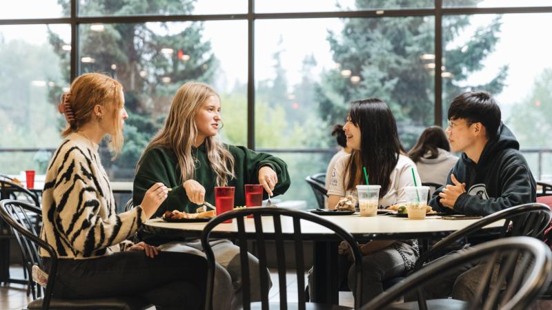 Four students sitting in cafeteria eating, smiling, talking.