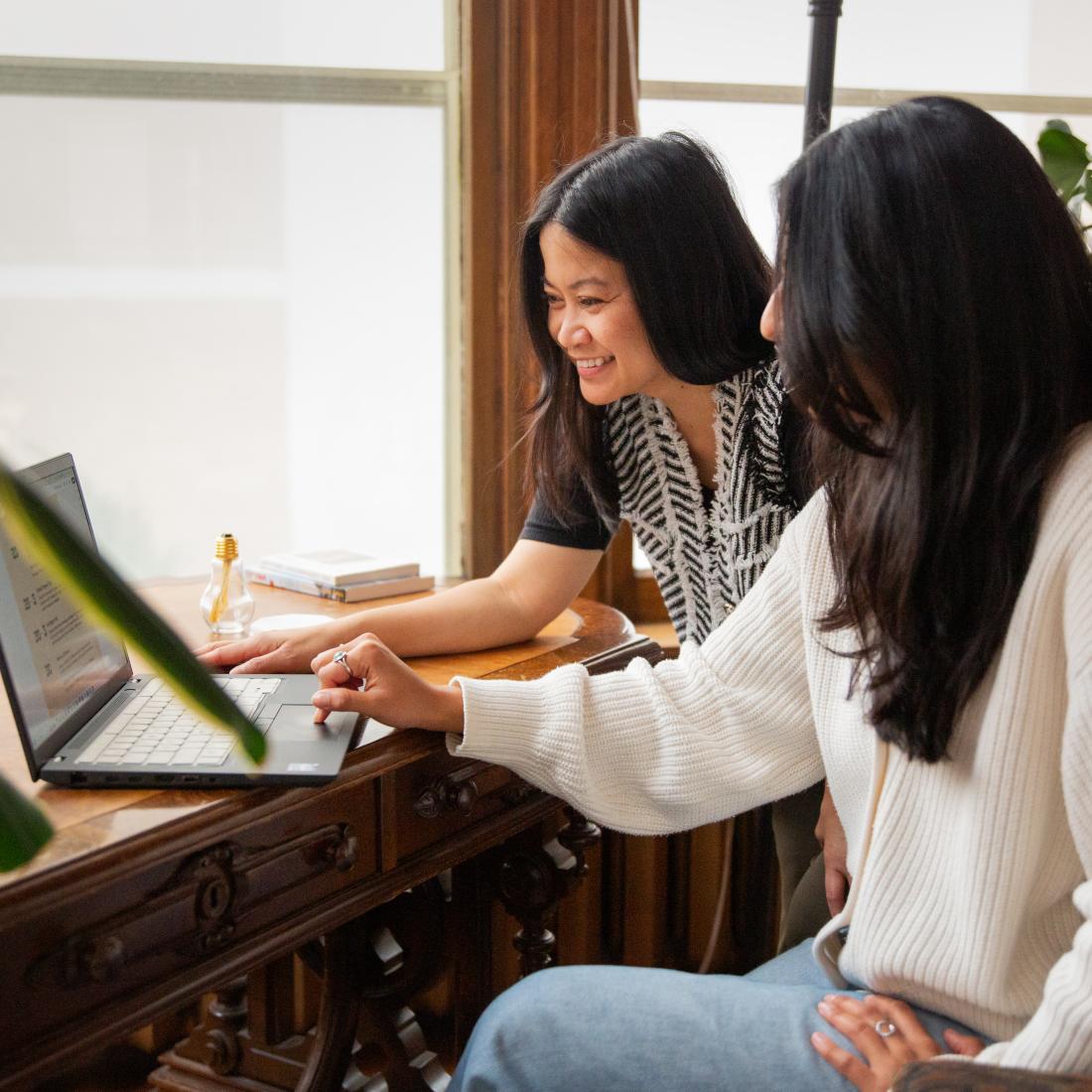 Two women sit at a wooden desk surrounded by plants and books in a bright room with large windows.