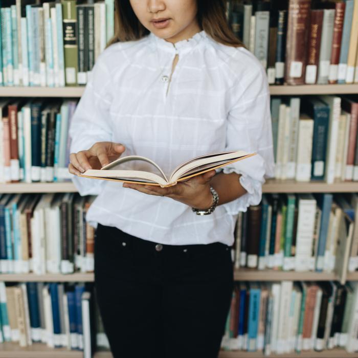 Student holding an open book in the library in front  of a shelf of books.