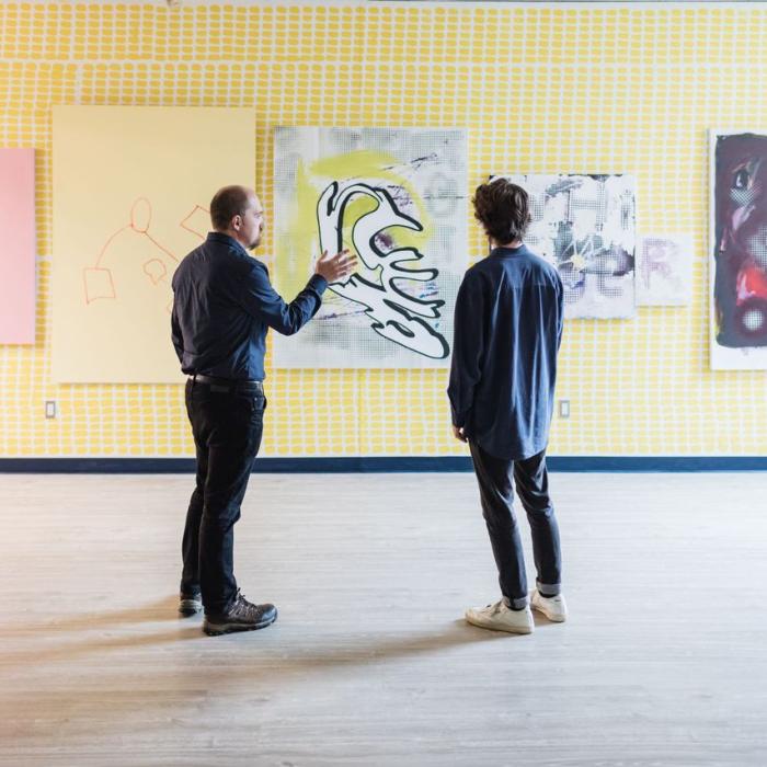 A professor and a student talking while looking at a wall displaying five art pieces.