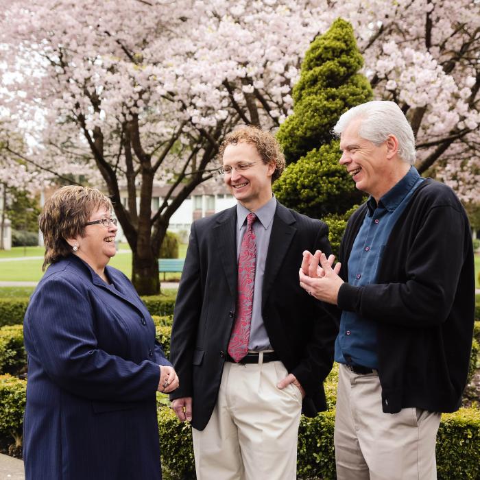 Three people talking outside with greenery and cherry blossom trees