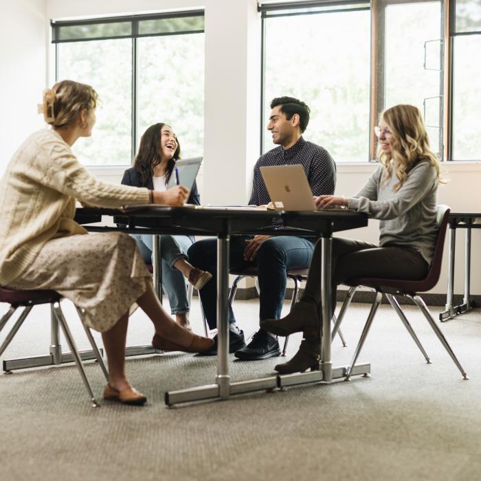 Four students sitting at a table studying on computers and laughing