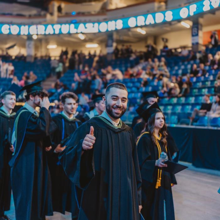 A group of graduates from Trinity Western University walk in a procession during the Spring Graduation Ceremony, wearing black academic gowns and hoods. A young man in the foreground smiles and gives a thumbs-up to the camera. Other graduates, some holding diplomas or programs, walk behind him. The venue is a large indoor arena with blue stadium seating, and a digital screen above displays "CONGRATULATIONS GRADS OF 2024." Family, friends, and faculty members are seated in the background