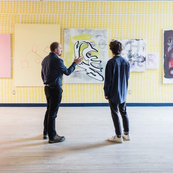 Two individuals stand in front of a wall with five large abstract paintings. The wall has a yellow checkered pattern, and the floor is wooden. They appear to be discussing the artwork.