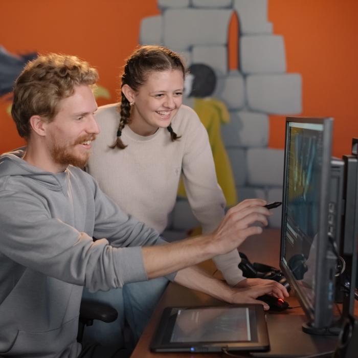 Two individuals work together at a computer workstation. One person uses a stylus on a graphics tablet, while the other points at the computer screen. The background features an orange wall with a mural of a dragon and stone structures.