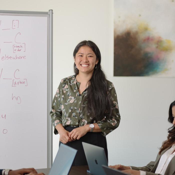 A person with long dark hair, wearing a green floral blouse, stands in front of a whiteboard and speaks to three seated individuals who are using laptops. The whiteboard contains partially visible handwritten notes and equations. A colourful abstract painting hangs on the wall in the background.