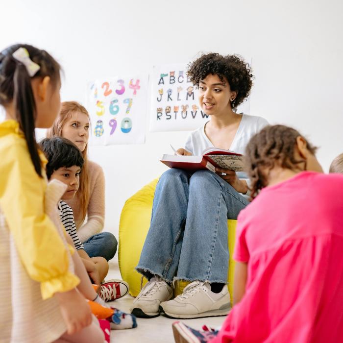 A teacher is sitting on a yellow bean bag chair, reading a book to a group of young children. The children are seated on red polka-dotted cushions around the teacher. On the wall behind them, there are posters with colourful numbers and letters.