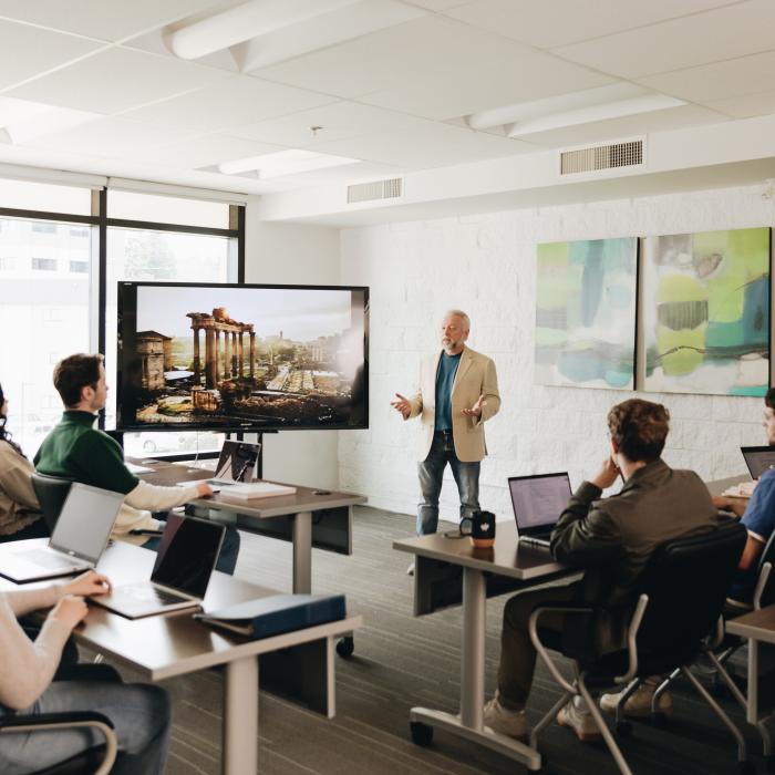 A group of university students seated with laptops in a classroom setting listen to a professor in front of a screen showing ancient ruins.