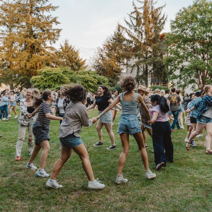 A group of people holding hands and dancing in a circle on a grassy field during an outdoor event.