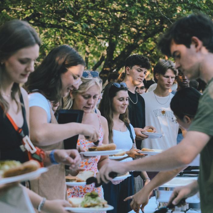 Young men and women standing in line at an outdoor buffet, serving themselves food in a park-like setting with trees in the background.
