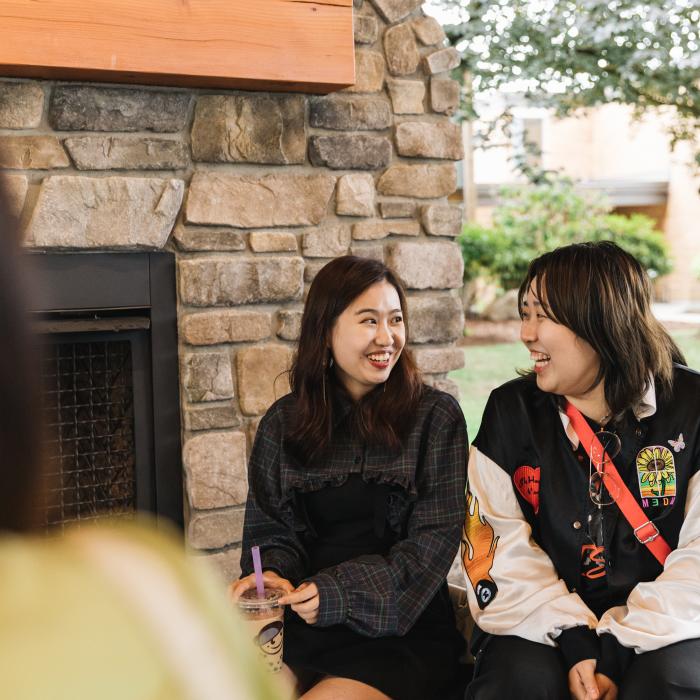 Two young women sit in front of a stone fireplace, smiling at each other. One holds a boba drink with a purple straw. A third person is partially visible in the foreground.