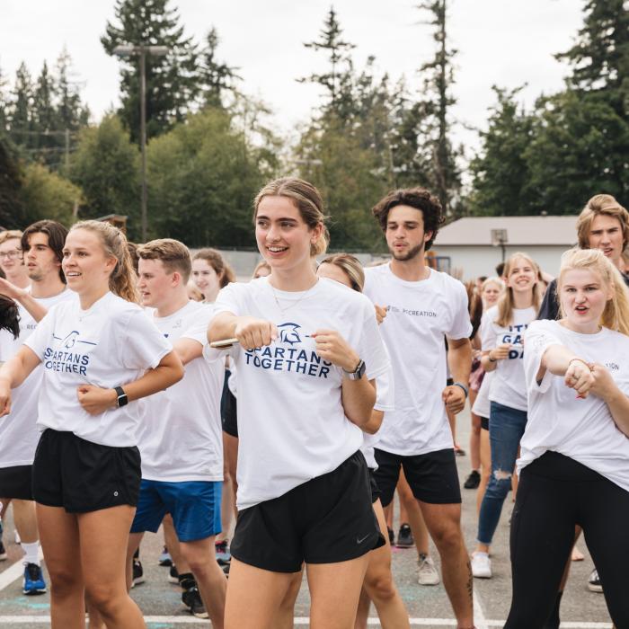 A group of young adults in white "SPARTANS TOGETHER" t-shirts participate in a synchronized outdoor activity, with trees and a building in the background.