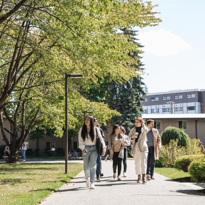 A group of people walk along a pathway in a campus-like setting, surrounded by trees, grass, and buildings.