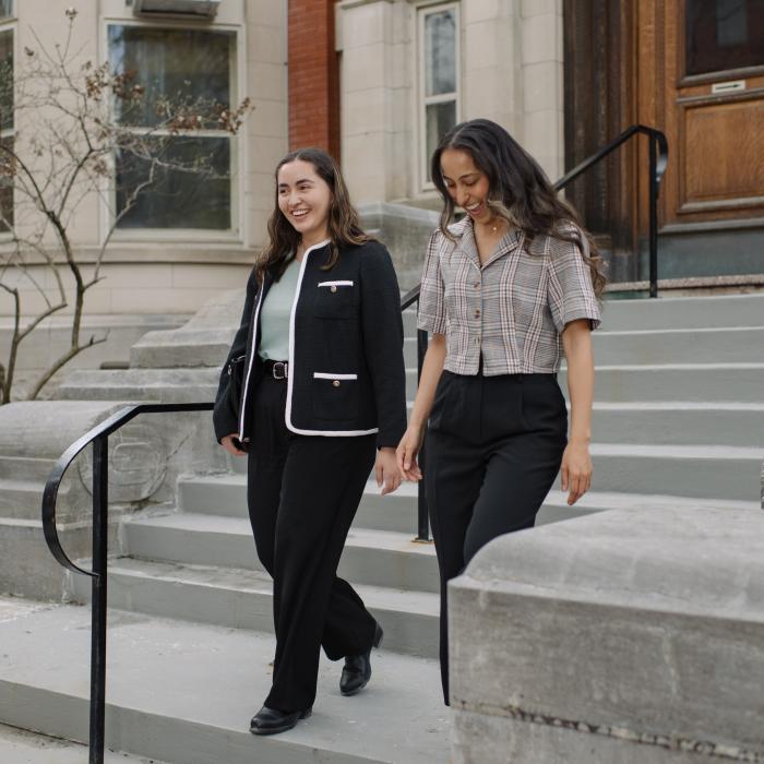 Two young women walk down stone steps outside a building with large windows and a wooden door, dressed in business casual attire.