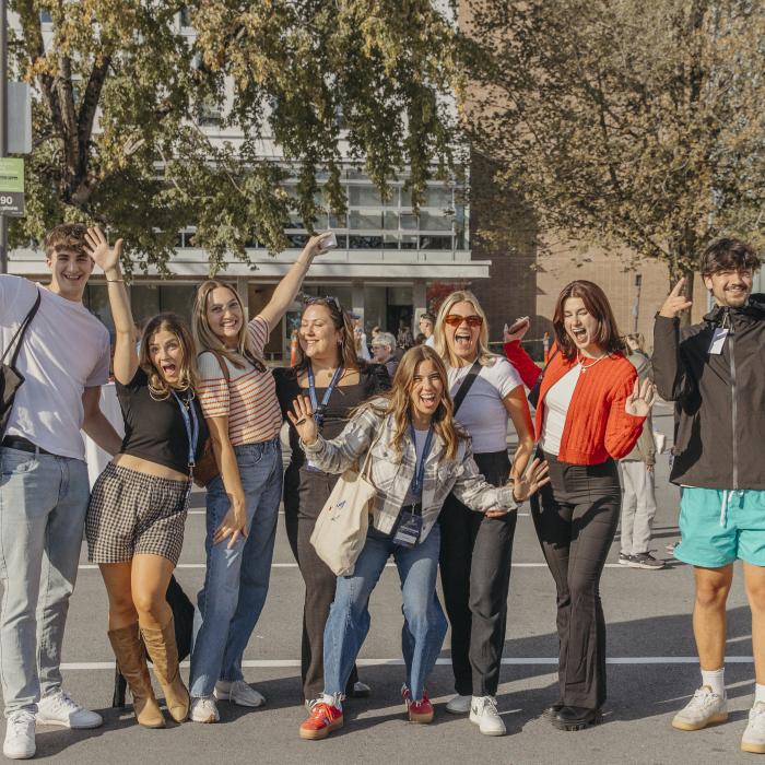 Group of nine young men and women posing casually in a parking lot with trees and a building in the background.