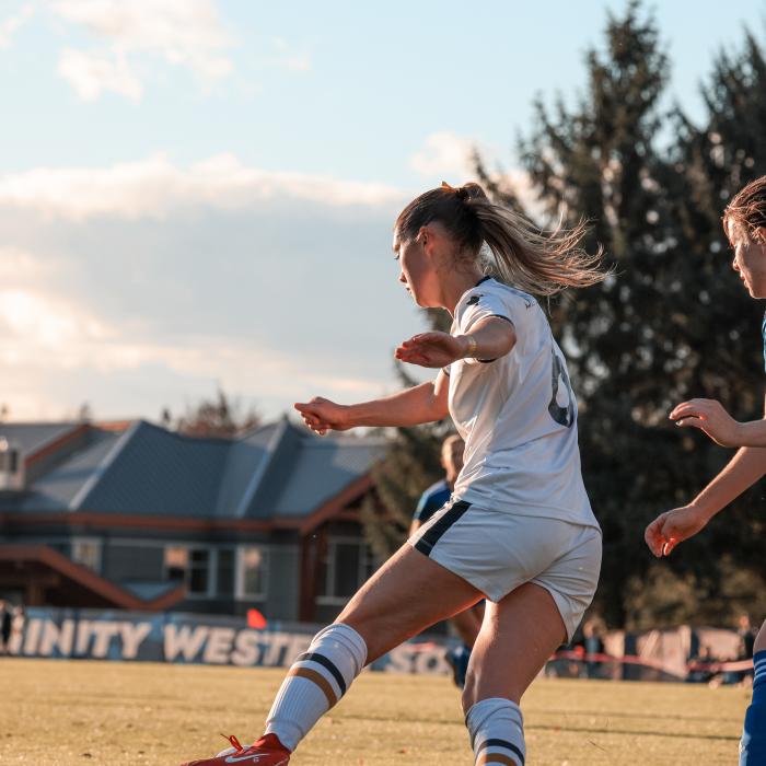 Two soccer players, one in white and one in blue, compete for the ball on an outdoor field, with a referee and teammates in the background. A "TRINITY WEST" banner is visible on the sideline.