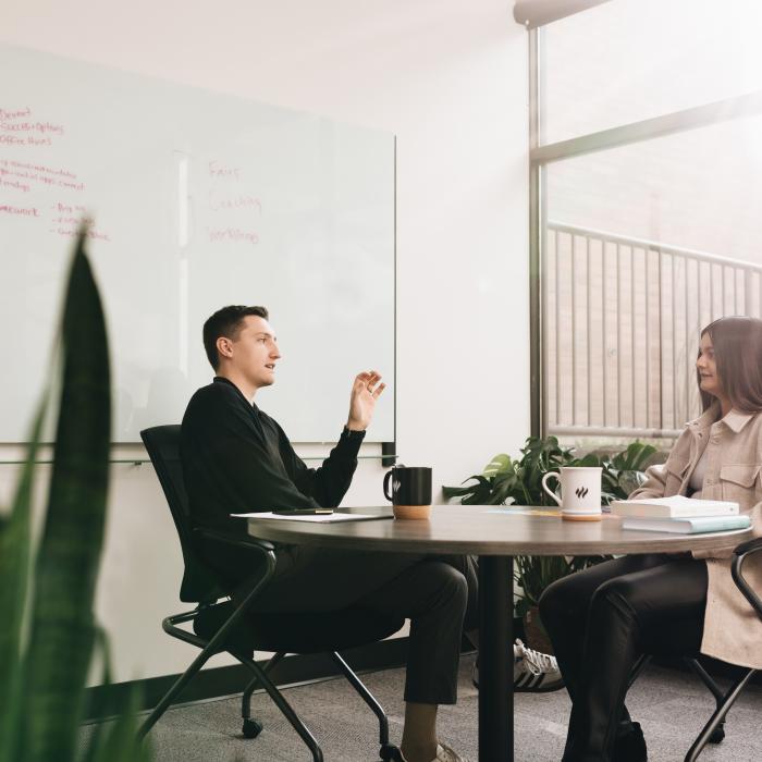 Two people sit at a round table in an office, talking, with a whiteboard behind them.