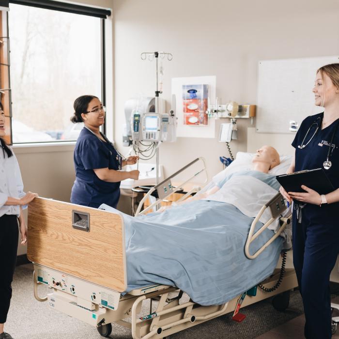 Three people stand around a patient in a hospital bed, surrounded by medical equipment and natural light from nearby windows.
