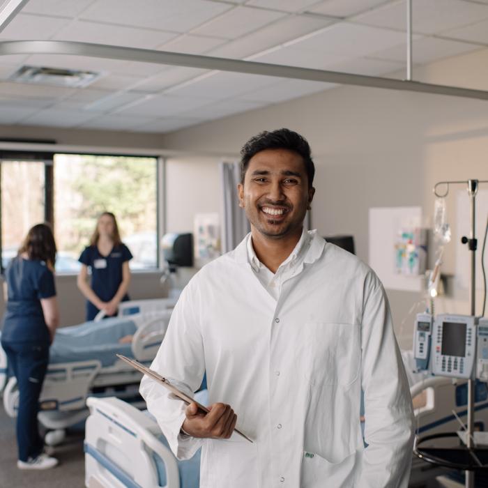 A person in a white lab coat holds a clipboard in a hospital room, while two others stand near a bed surrounded by medical equipment.