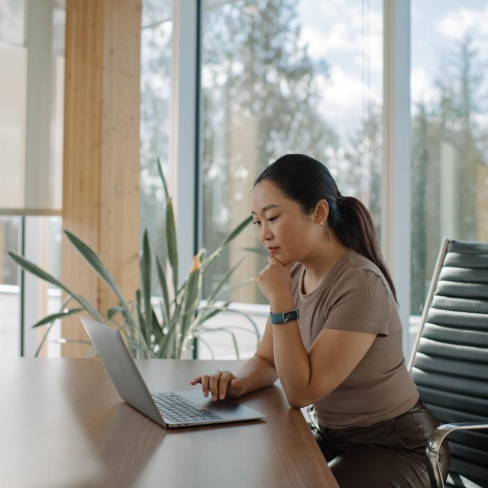 A young woman sits at a desk in front of a laptop, one hand on the keyboard and the other resting on their chin.