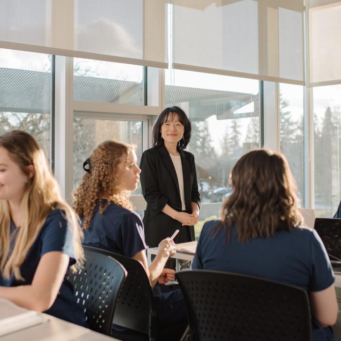 A group of students sit around a classroom table with notebooks and laptops while an instructor stands at the head of the table.