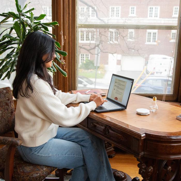  Person in a white sweater working on a laptop at a wooden desk near large windows with a city view.
