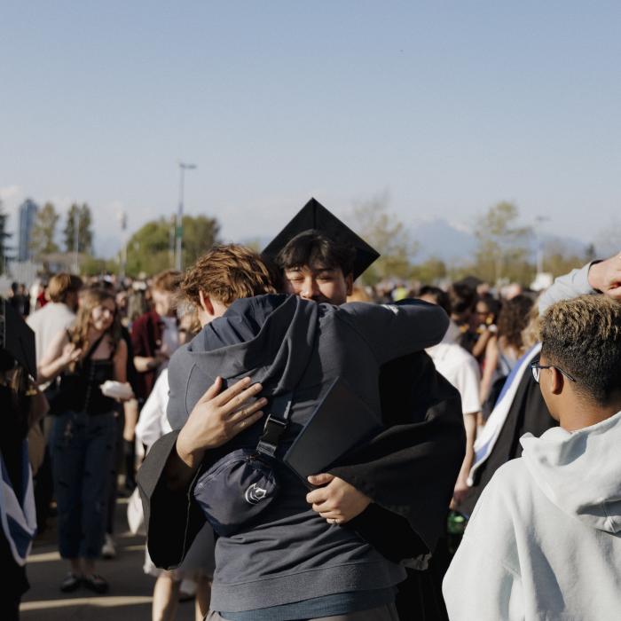 A person in a graduation cap and gown hugs someone during an outdoor ceremony, surrounded by people with flowers and gifts, with trees and mountains in the background.