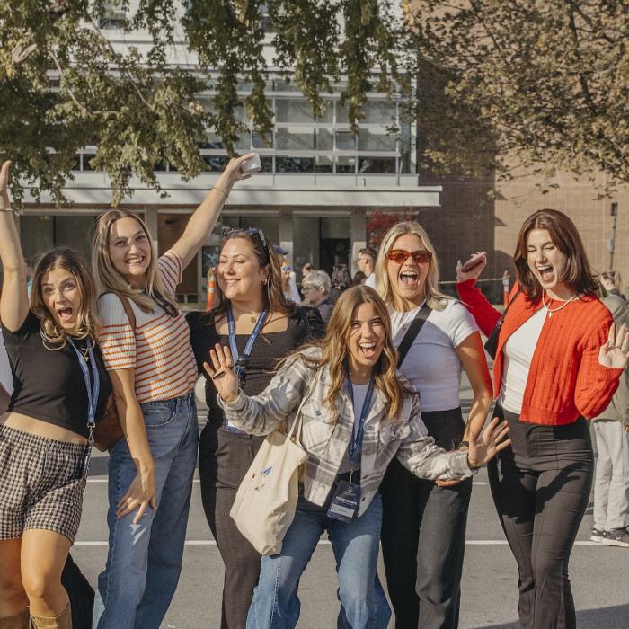 Group of nine young men and women posing casually in a parking lot with trees and a building in the background.