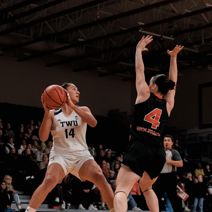 TWU basketball player in white jersey number 14 holds the ball while being closely defended by a player in black jersey number 4.