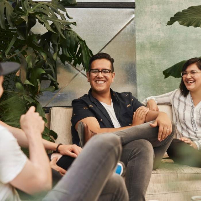 Three people laughing and chatting on a sofa surrounded by plants.